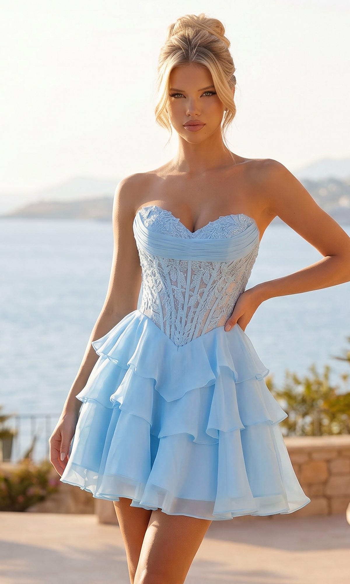Wearing the PromGirl Exclusive 250334, a light blue strapless short homecoming dress with a delicate lace bodice, a woman stands outdoors by a scenic waterfront with majestic mountains in the background.